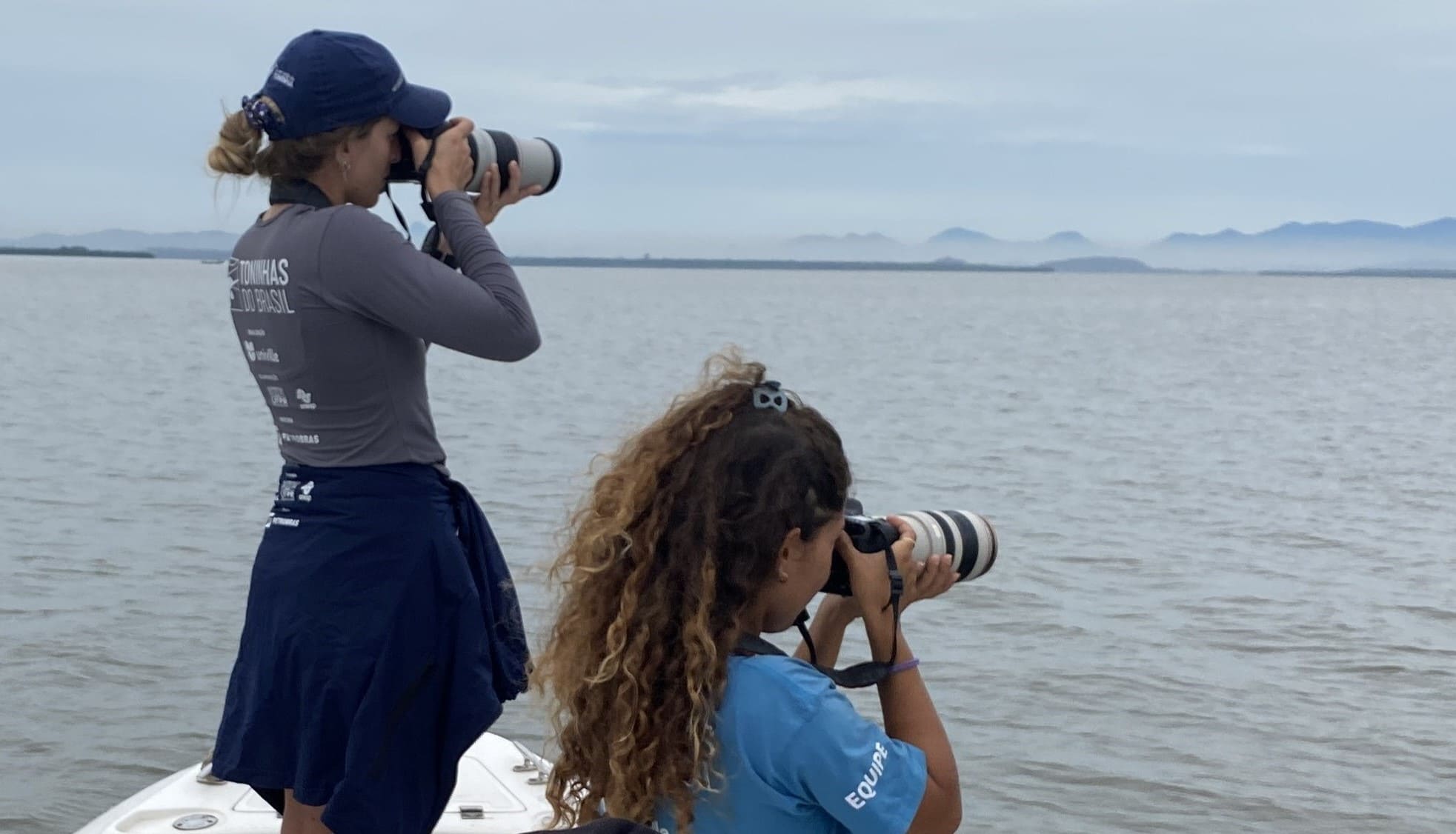 Duas pesquisadoras do Toninhas do Brasil empunhando câmeras fotográficas enquanto monitoram as toninhas da Baía Babitonga.